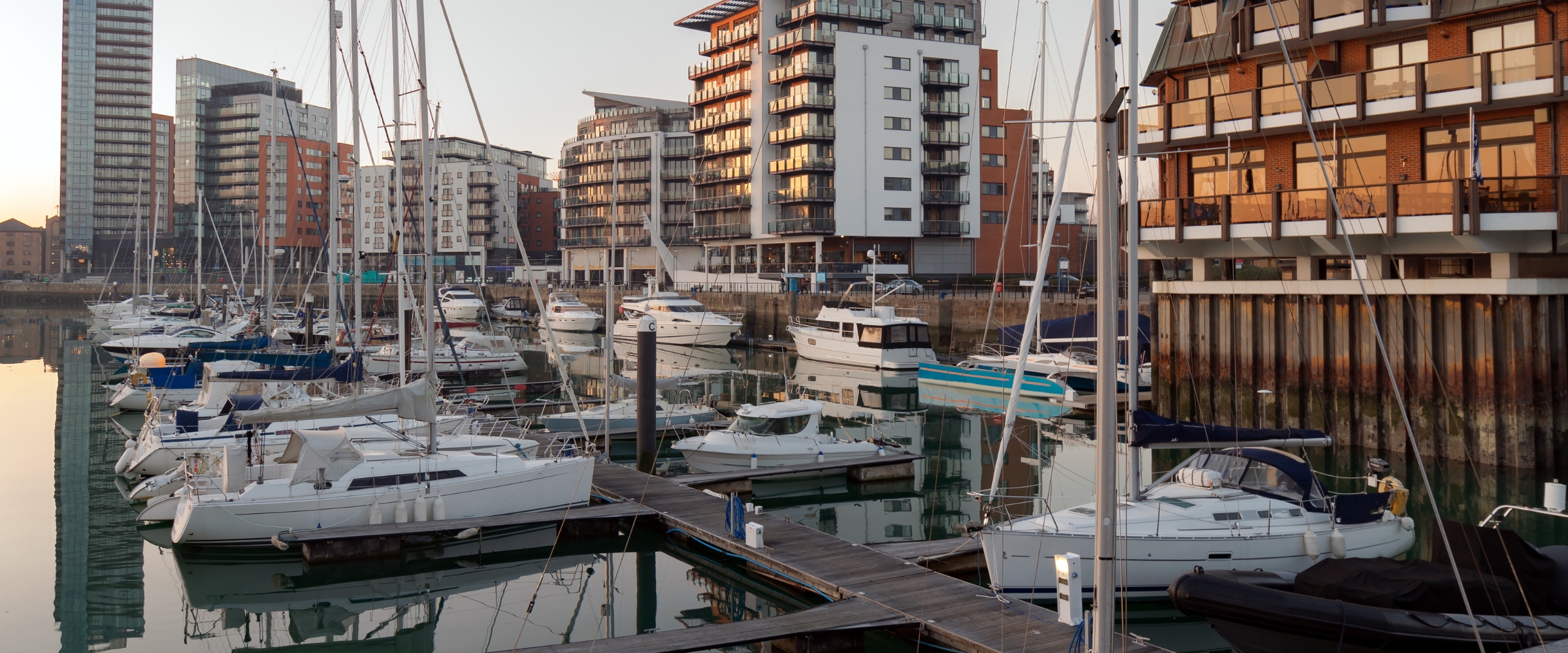 Yachts moored in a city marina at dusk, with apartment buildings along the quay.